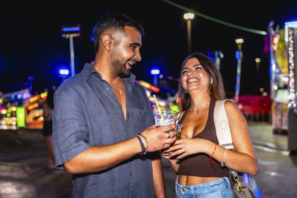 Young man and woman laughing joyfully while enjoying cocktails together, surrounded by the vibrant