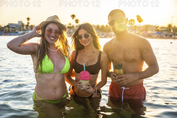 Three friends enjoying refreshing cocktails while standing in ocean water during a vibrant sunset