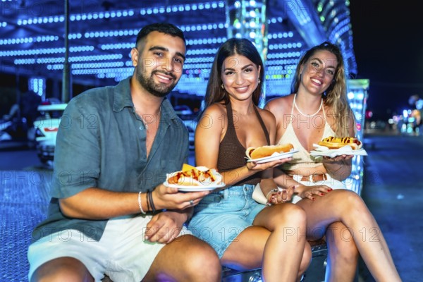 Three friends are sitting and smiling, holding hot dogs on paper plates at a brightly lit amusement