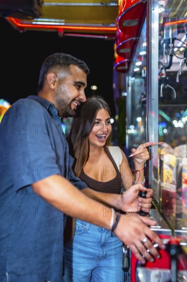 Young couple having fun playing a claw crane game at an amusement park at night, trying to win a