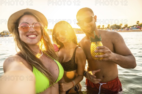 Three cheerful friends are taking a selfie while having a refreshing cocktail in the sea during a