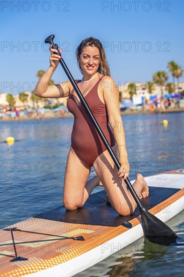 Woman kneeling on a paddleboard, enjoying a sunny day at the beach. Clear blue sky and palm trees