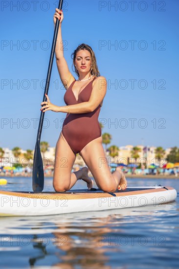 Young woman in a swimsuit kneels on a paddle board, holding a paddle and enjoying a summer day on