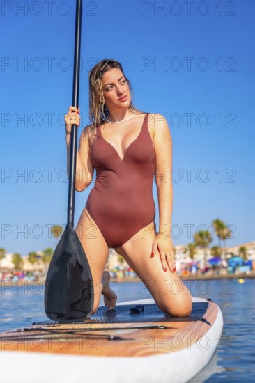 Young woman kneeling on paddle board, holding paddle and looking away, enjoying summer sunny day at