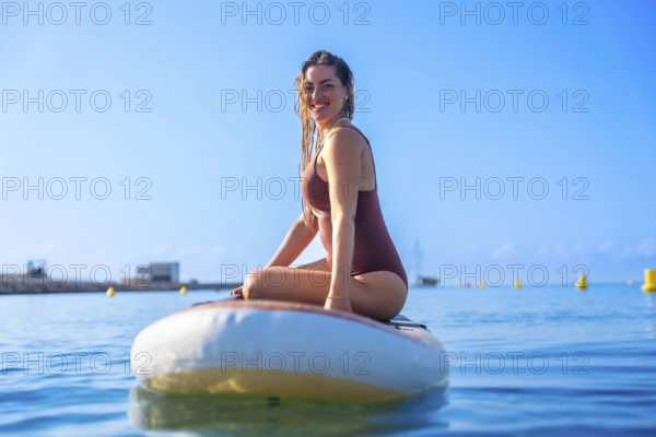 Smiling woman sitting on a paddleboard in clear blue waters, enjoying a sunny summer day. Perfect
