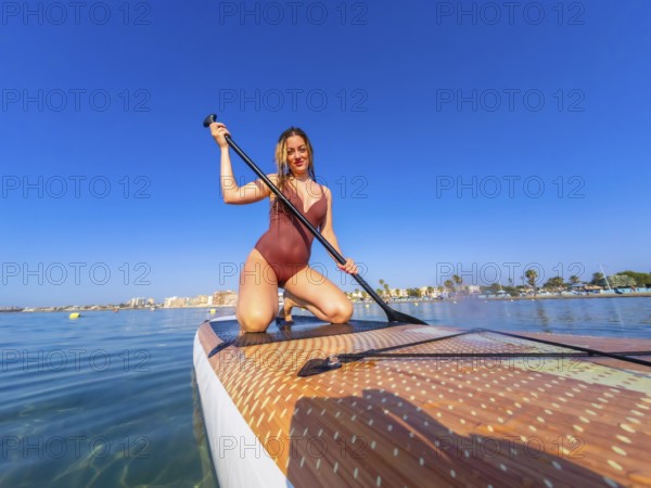 Young woman kneeling on a paddle board, holding a paddle, enjoying a summer watersport activity on