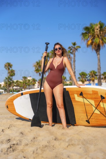 Young woman in swimsuit holding a paddle and posing with her paddle board on the beach on a sunny