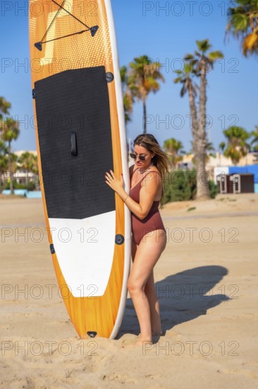 Young woman holding a paddle board on a sandy beach with palm trees in the background, enjoying
