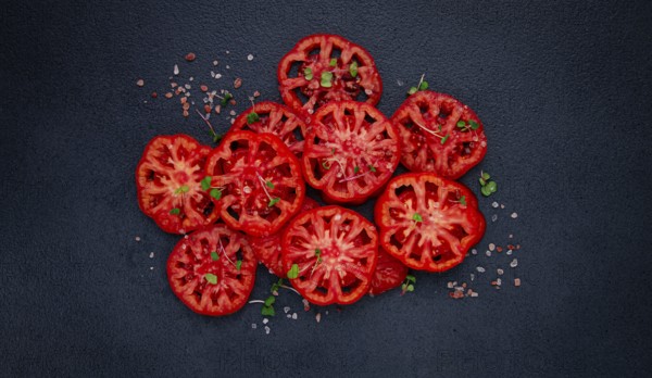 Freshly sliced ripe tomatoes arranged on a textured dark surface with herbs and salt scattered