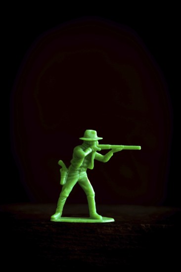 A vibrant green plastic toy cowboy stands in a tactical pose, aiming a rifle against a dark