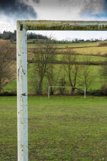 A weathered soccer goalpost stands in the foreground of a wide grassy field, with lush green hills