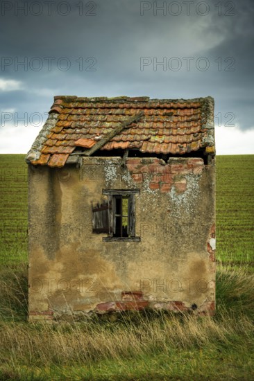 A small, dilapidated hut with a broken roof and cracked walls sits alone in a vast green field.
