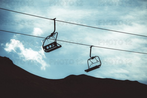 Two empty ski lifts hang from cables, silhouetted against a backdrop of a dark mountain and cloudy