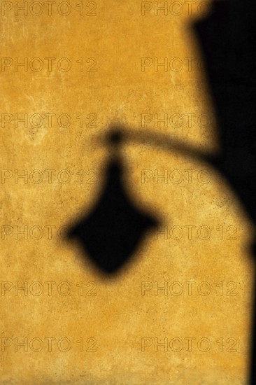 Soft afternoon light creates a striking shadow of a lantern on a textured yellow wall. The warm