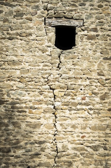 A weathered stone wall displays a prominent crack running vertically, featuring a small window.