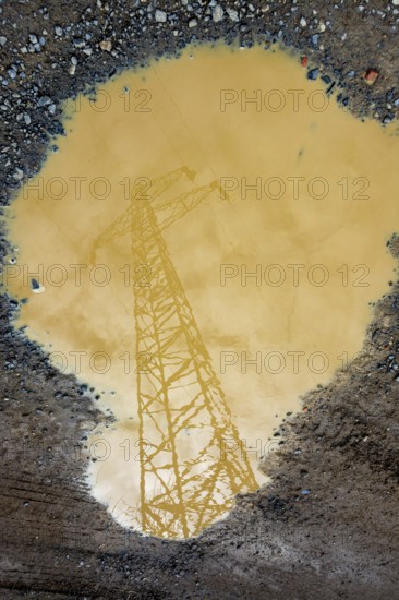 A power line tower is reflected in a large puddle of muddy water. The cloudy sky is visible, adding