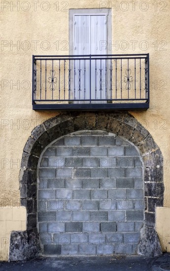 A metal-railed balcony stands over a blocked stone entrance on a textured yellow building, creating