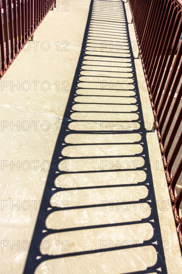 Bright sunlight creates intricate shadows from a red railing onto a stone pathway in a park,