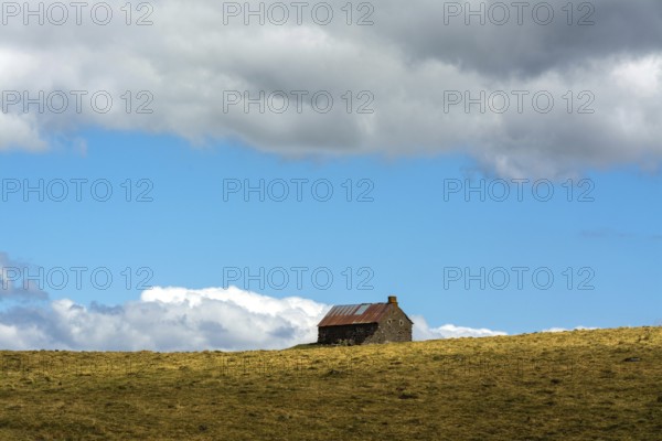 A stone cottage rests on a grassy slope, surrounded by expansive hills. The sky features vibrant