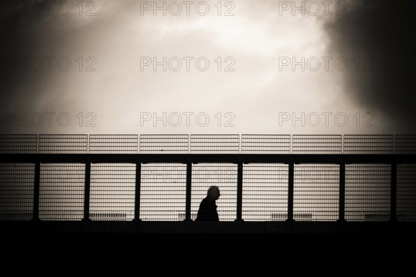 A solitary figure walks along a metal gangway in Lausanne, Switzerland, with a dramatic cloudy sky