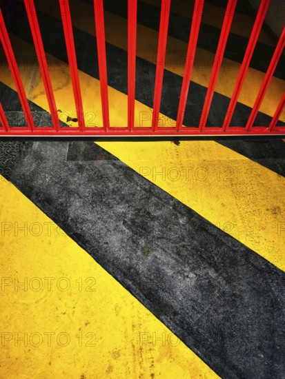 Bright yellow and black stripes intersect with a bold red barrier across a concrete surface. This