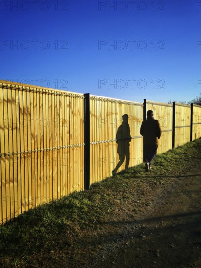 A solitary figure strolls beside a tall wooden fence on a well-worn path. The sun casts elongated