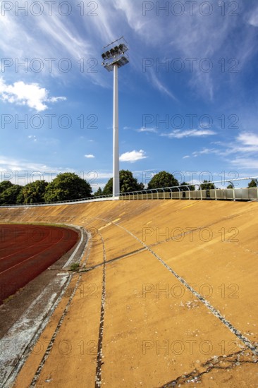 Bright daylight reveals an empty sports stadium with a vibrant red track. The tall floodlights,