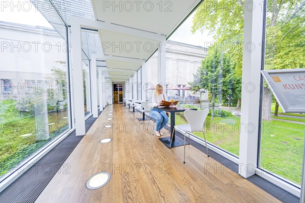 Woman sitting in a bright café with glass walls and a green garden view, Frieder Burda Museum,