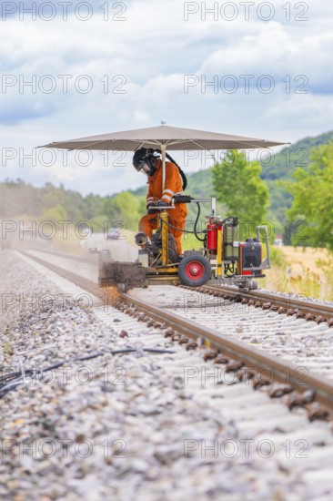 Worker on railway tracks uses a machine under a screen. Dust and nature in the background, grinding