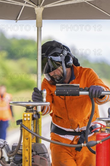 A worker in an orange suit and helmet works under an umbrella on a railway line, grinding work on