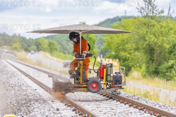 Worker in orange overalls operating a machine on railway tracks, protected by a large umbrella,