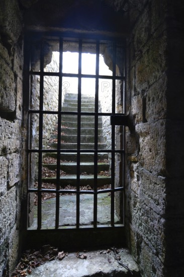 A medieval dungeon in France features thick stone walls and a barred window revealing illuminated