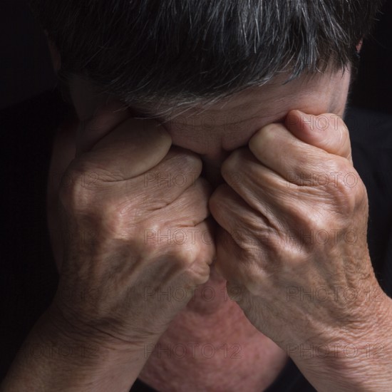 A person sits with their head bowed, hands clasped tightly over their eyes, conveying deep emotion