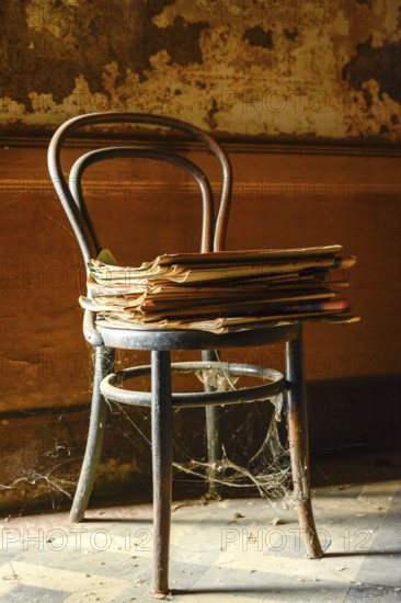 An old wooden chair is prominently positioned with a pile of yellowed newspapers resting on the