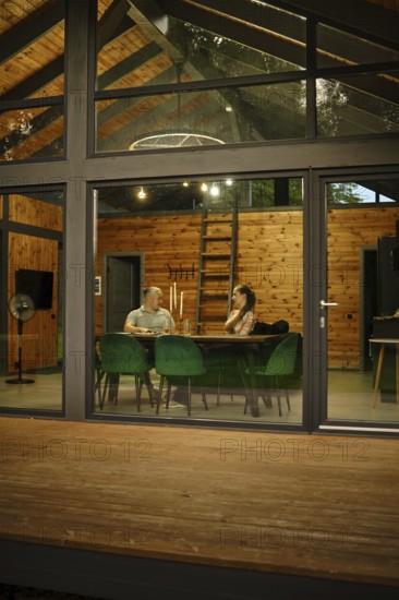 A modern wooden cabin, viewed from outside in the evening. Through large glass windows and doors a