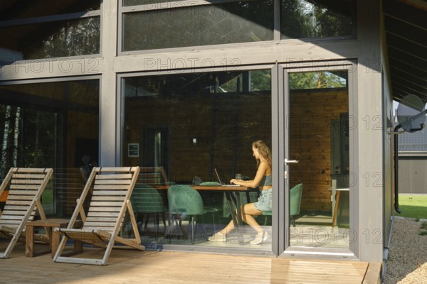 A woman is inside a modern country house with large windows and watches a TV series on a laptop