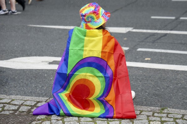 A participant with a rainbow flag and colourful hat sits on the side of the road at the 47th