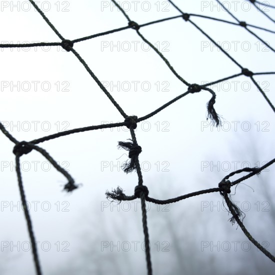 A close-up view of worn netting with frayed edges, suspended in a misty landscape. The atmosphere