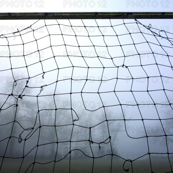 A weathered football goal net looms in the foreground surrounded by dense fog, with a blurred view