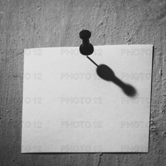 A blank piece of note paper is pinned to a textured wall, casting a compelling shadow that