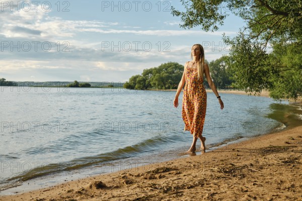 A woman in a vibrant sundress strolls barefoot along the sandy beach beside a calm lake. Sunshine