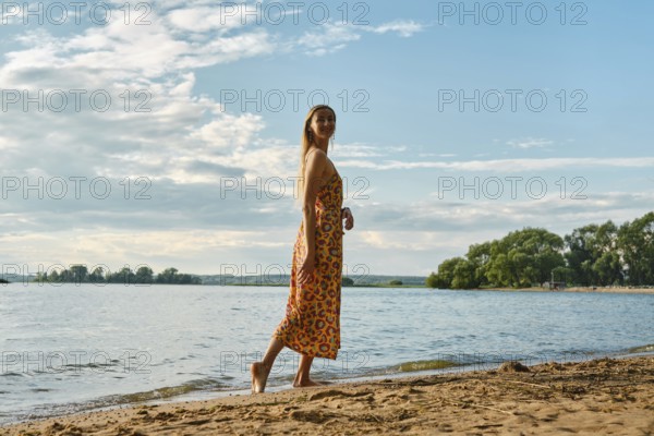 A woman is strolling barefoot along a sandy riverbank, wearing a colourful floral sundress. The