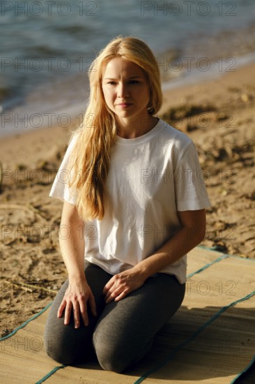 A young woman sits cross-legged on a mat at the beach, gazing thoughtfully towards the water. The