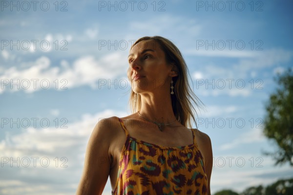 A woman stands outdoors, wearing a vibrant, colourful dress. She gazes into the distance as the sun
