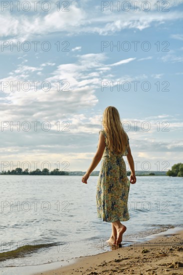 Rear view of barefoot woman standing on a sandy beach by a tranquil lake. The clear blue sky