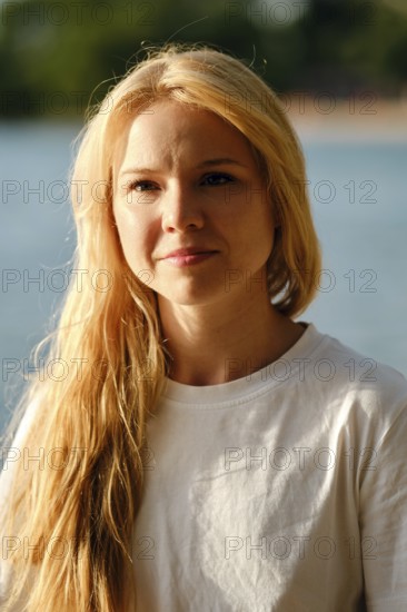 A woman with long flowing hair stands by a serene body of water. Her relaxed expression showcases