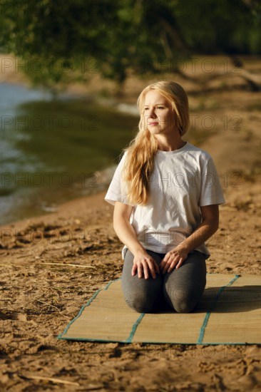 A woman sits on a mat by the riverside, engaged in a moment of mindfulness. She has long blonde