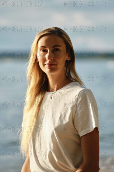 A woman with long hair smiles gently while standing near the water's edge at the beach. The bright