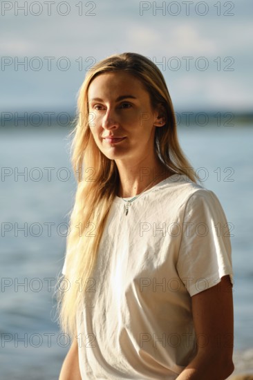 A woman with long blonde hair is standing near the water's edge at sunset. She gazes thoughtfully