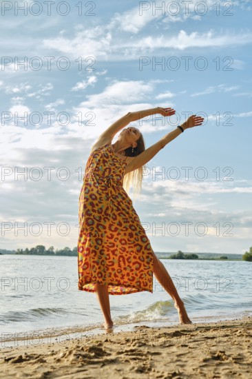A joyful woman stretches gracefully on the sandy shore as the sun sets over the water. The vibrant